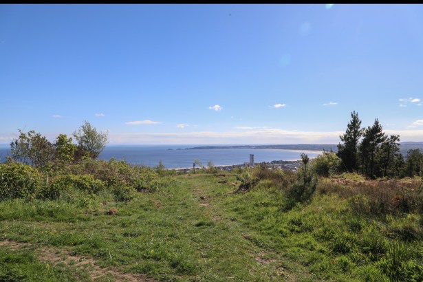 View from the grassy summit of Kilvey Hill over Swansea city centre and the curve of Swansea Bay towards Mumbles under a blue sky