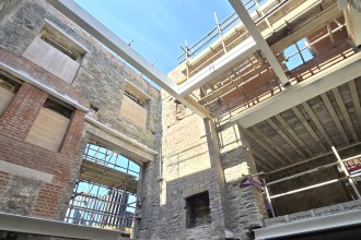 Steel beams and timber joists span the interior of the roofless stone Laboratory Building, with scaffolding and blue sky above