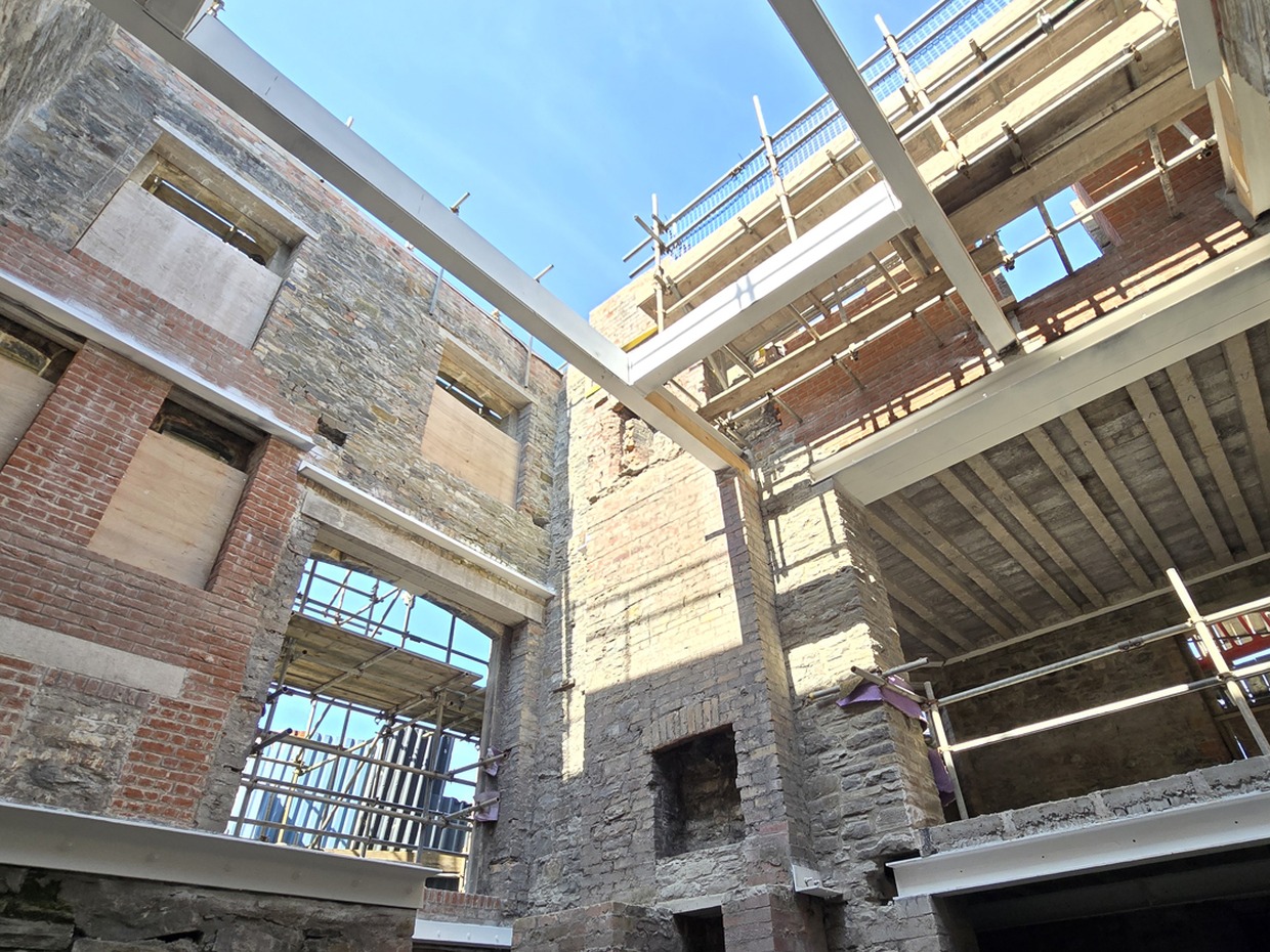 Steel beams and timber joists span the interior of the roofless stone Laboratory Building, with scaffolding and blue sky above
