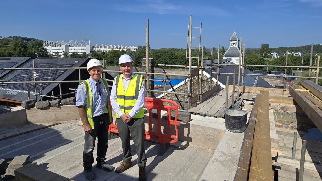 Two men in hard hats and hi-vis vests stand on the roof of the Laboratory Building with the Swansea.com Stadium and a clock tower behind