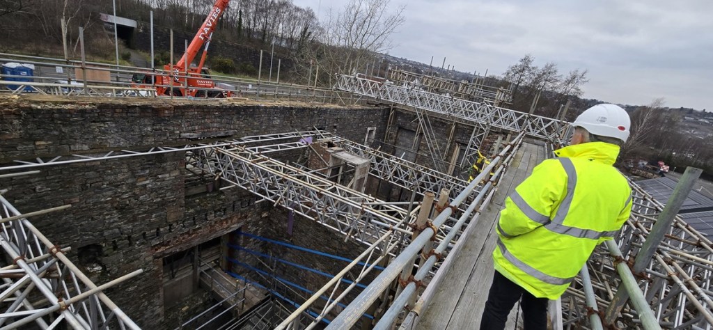 A worker in hi-vis on scaffolding looks down into the roofless stone interior of the Laboratory Building, with an orange crane behind