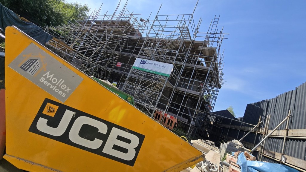 The Laboratory Building encased in scaffolding, seen past a yellow JCB, with a John Weaver Conservation banner on the frame