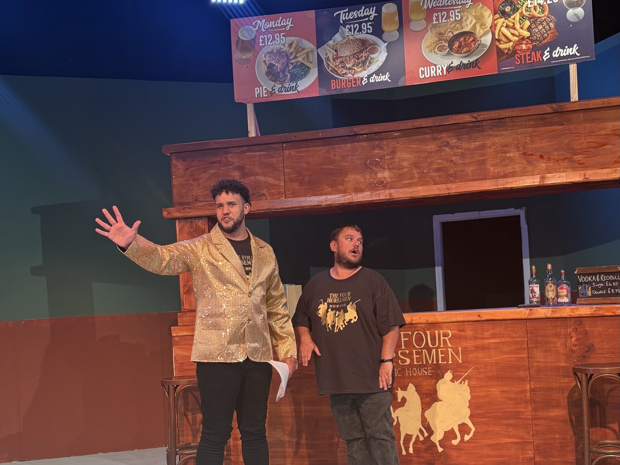 Dylan Fairbairn in a gold sequinned jacket and John Welch on stage in front of the pub set with a meal deal banner above