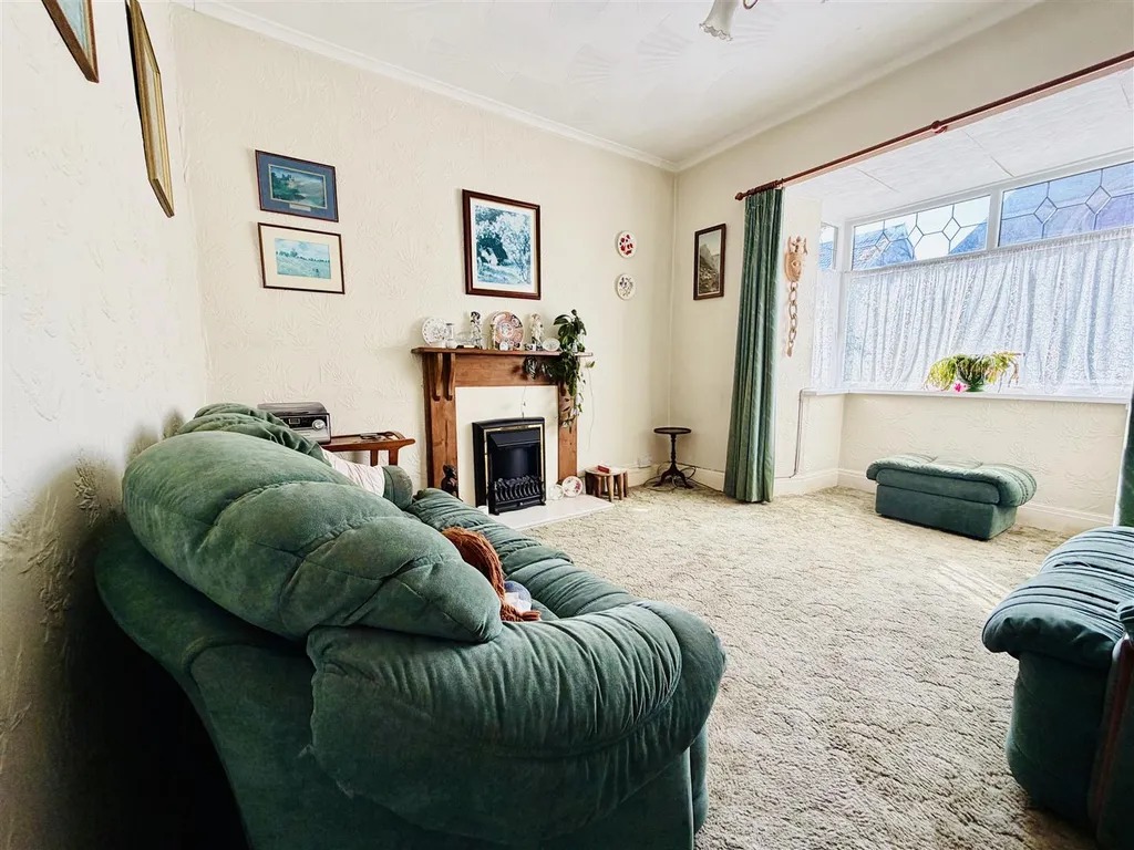 A lounge with a green three piece suite, a bay window in lace curtains and a fireplace
