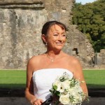 Lynne Trump smiling in a white wedding dress and holding a bouquet of white flowers in front of stone abbey ruins