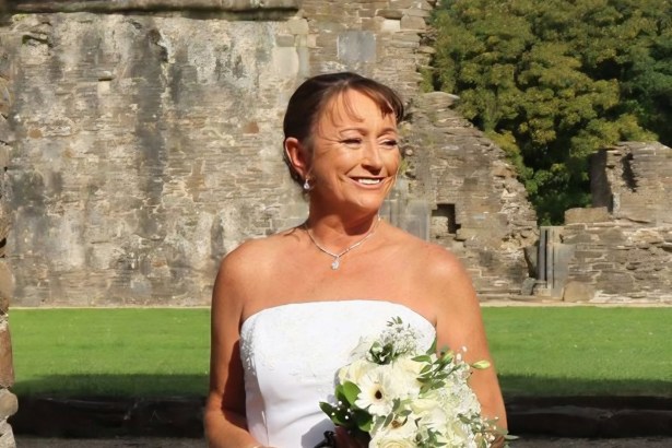 Lynne Trump smiling in a white wedding dress and holding a bouquet of white flowers in front of stone abbey ruins