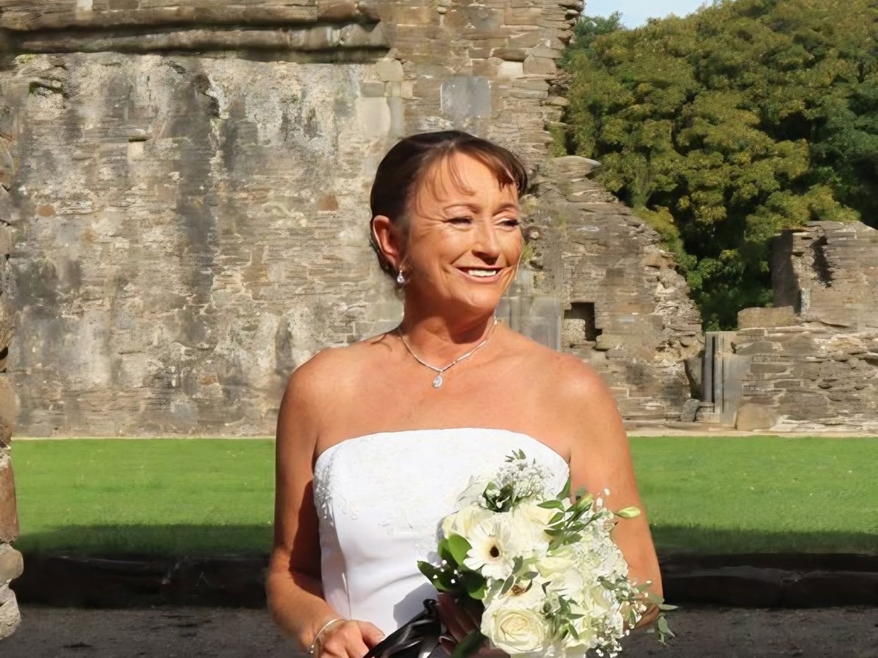 Lynne Trump smiling in a white wedding dress and holding a bouquet of white flowers in front of stone abbey ruins