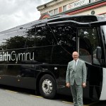 Deputy Minister for Transport Mark Hooper in a grey suit standing in front of a black TaithCymru coach outside Bangor railway station