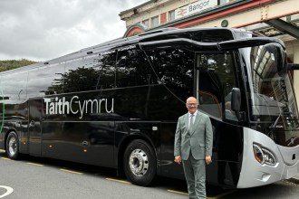 Deputy Minister for Transport Mark Hooper in a grey suit standing in front of a black TaithCymru coach outside Bangor railway station