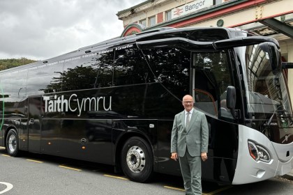 Deputy Minister for Transport Mark Hooper in a grey suit standing in front of a black TaithCymru coach outside Bangor railway station