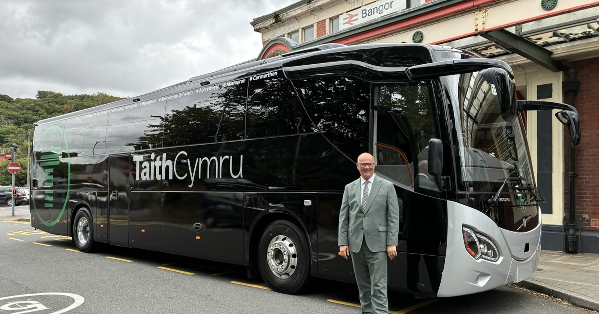 Deputy Minister for Transport Mark Hooper in a grey suit standing in front of a black TaithCymru coach outside Bangor railway station