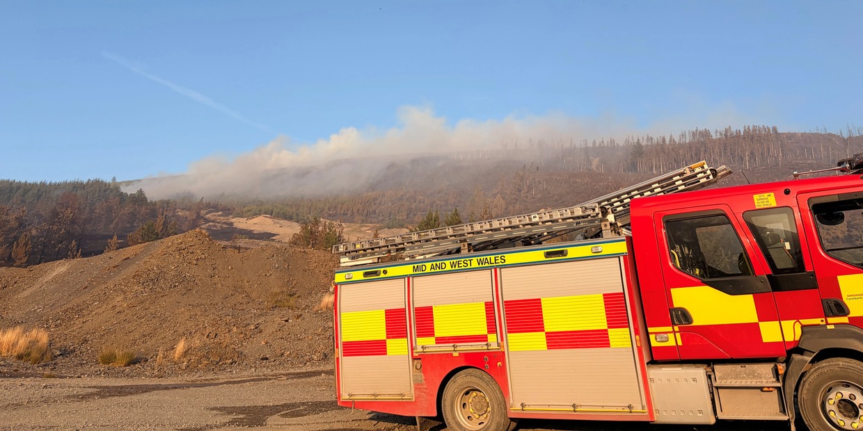 A Mid and West Wales Fire and Rescue Service fire engine parked beneath a scorched, smoking forested hillside under a blue sky