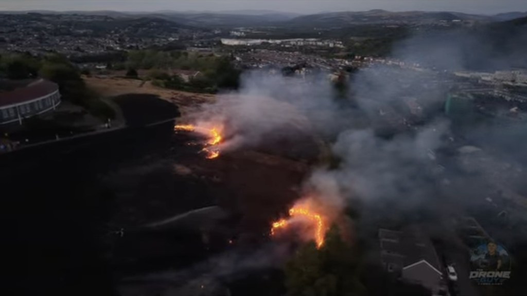 Aerial view of flames spreading across blackened hillside above Mayhill, with smoke rising over rooftops below