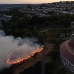 Aerial view of a line of flames and smoke burning across the hillside above Mayhill, with Seaview school and Swansea Bay beyond
