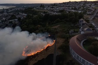 Aerial view of a line of flames and smoke burning across the hillside above Mayhill, with Seaview school and Swansea Bay beyond