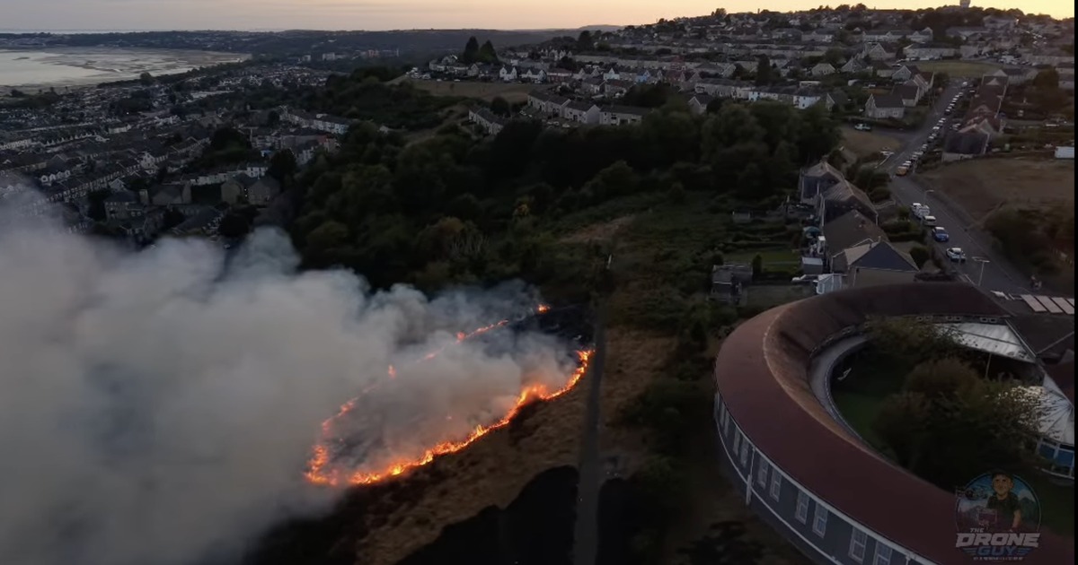 Aerial view of a line of flames and smoke burning across the hillside above Mayhill, with Seaview school and Swansea Bay beyond