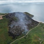 Aerial view of a large blackened burn scar covering a coastal headland above a beach, smoke drifting and fire engines on a track below