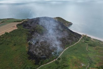 Aerial view of a large blackened burn scar covering a coastal headland above a beach, smoke drifting and fire engines on a track below