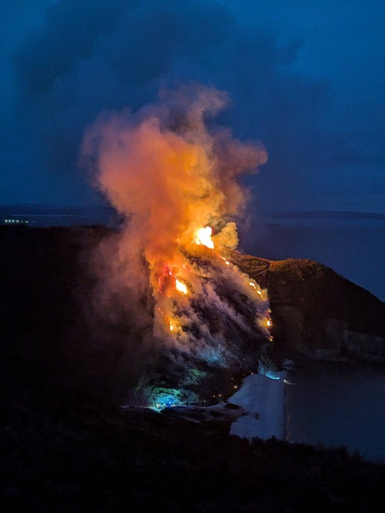 A wildfire burning down a steep coastal cliff towards a beach at night