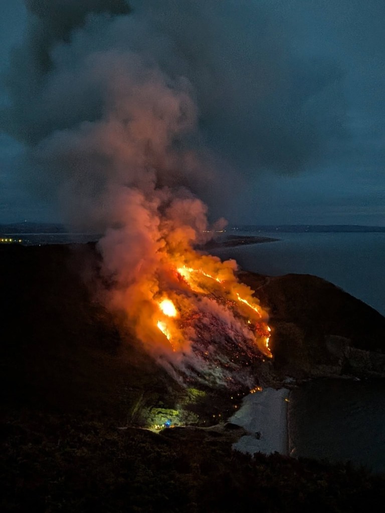 A burning hillside above Carmarthen Bay with heavy smoke rising at dusk