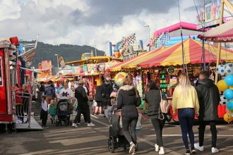 Crowds walking between stalls and rides at the Neath Great Fair funfair on Milland Road, with hills behind