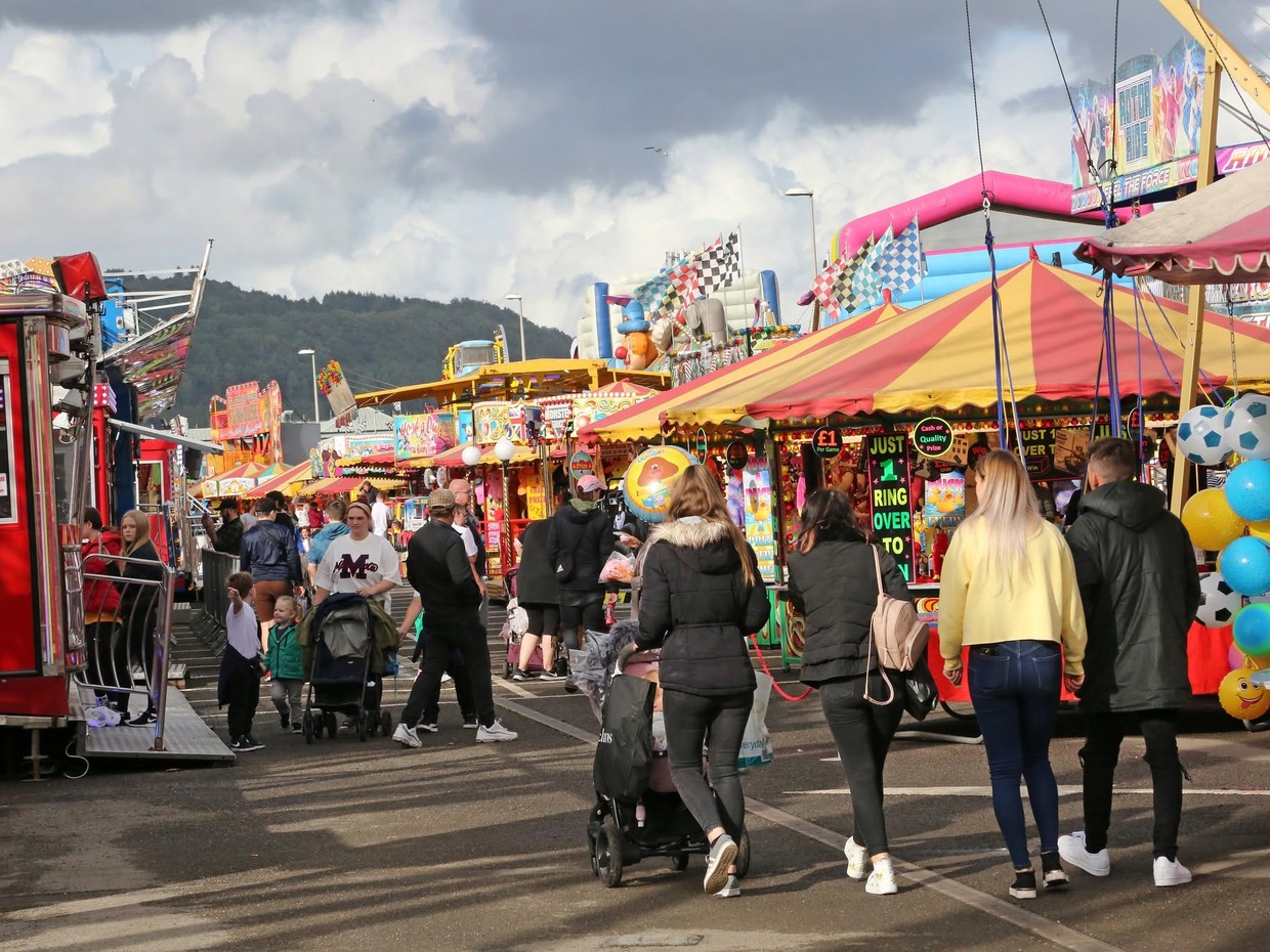 Crowds walking between stalls and rides at the Neath Great Fair funfair on Milland Road, with hills behind