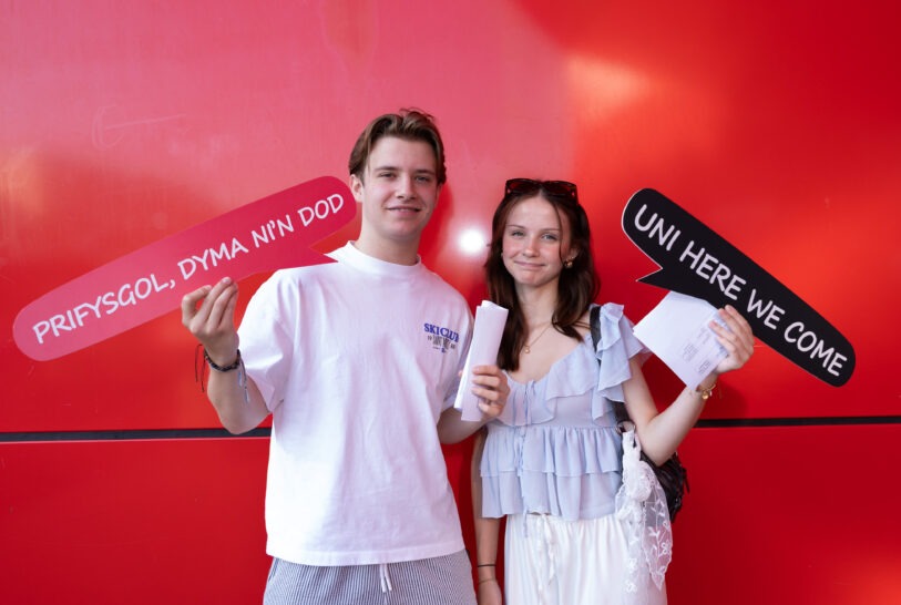 Two students hold bilingual Uni here we come and Prifysgol dyma ni'n dod signs against a red wall