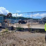 A Natural Resources Wales officer in hi-vis and a white hard hat looks through the fence at waste piles and machinery at the AMG Resources site in Llanelli