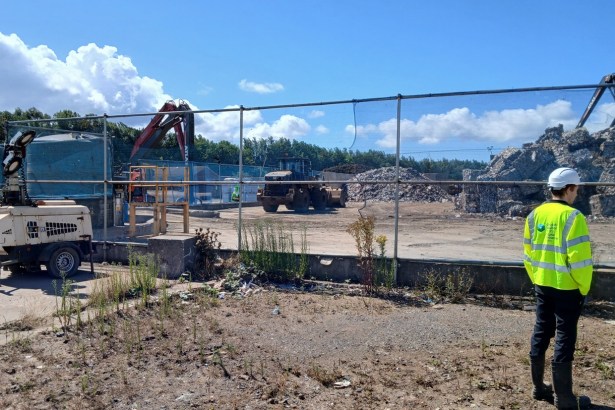 A Natural Resources Wales officer in hi-vis and a white hard hat looks through the fence at waste piles and machinery at the AMG Resources site in Llanelli