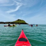 Sea kayakers paddling on calm turquoise water towards Worm's Head at Rhossili, seen from the bow of a red kayak
