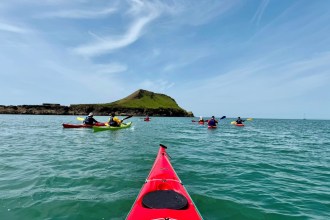Sea kayakers paddling on calm turquoise water towards Worm's Head at Rhossili, seen from the bow of a red kayak