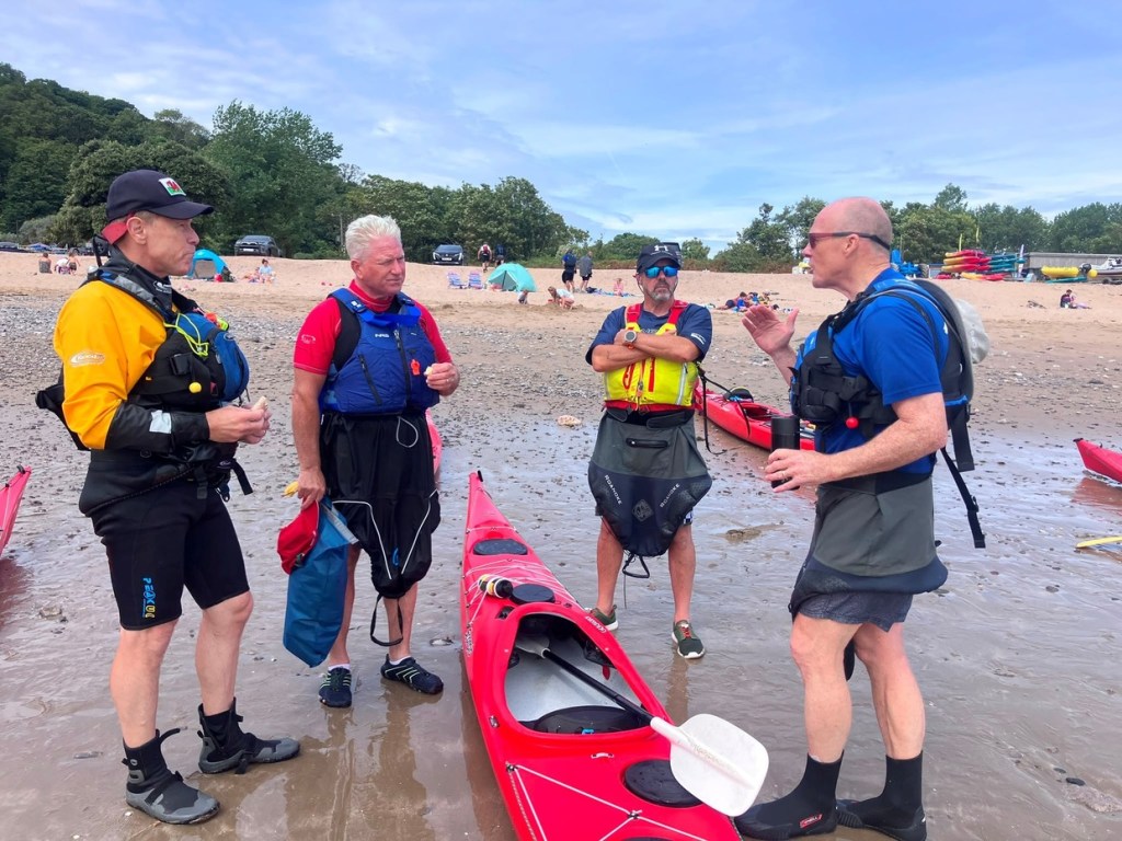 Four men in kayaking gear and buoyancy aids talking on a beach beside a red sea kayak