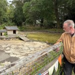A man in a tan jacket leans on a fence beside the drained duck pond at Parc Howard, with wooden shelters on an island in a dry cracked basin