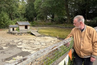 A man in a tan jacket leans on a fence beside the drained duck pond at Parc Howard, with wooden shelters on an island in a dry cracked basin