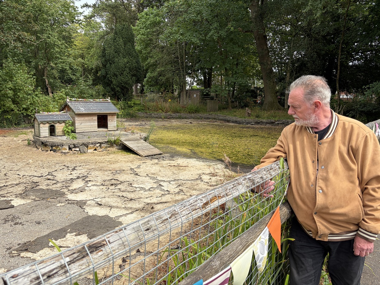 A man in a tan jacket leans on a fence beside the drained duck pond at Parc Howard, with wooden shelters on an island in a dry cracked basin