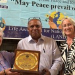 Pam Evans MBE, Mubarak Ali and Cllr Andrea Harrington holding the Leadership and Excellence Award plaque in front of a Peace Mala banner