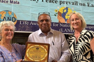 Pam Evans MBE, Mubarak Ali and Cllr Andrea Harrington holding the Leadership and Excellence Award plaque in front of a Peace Mala banner