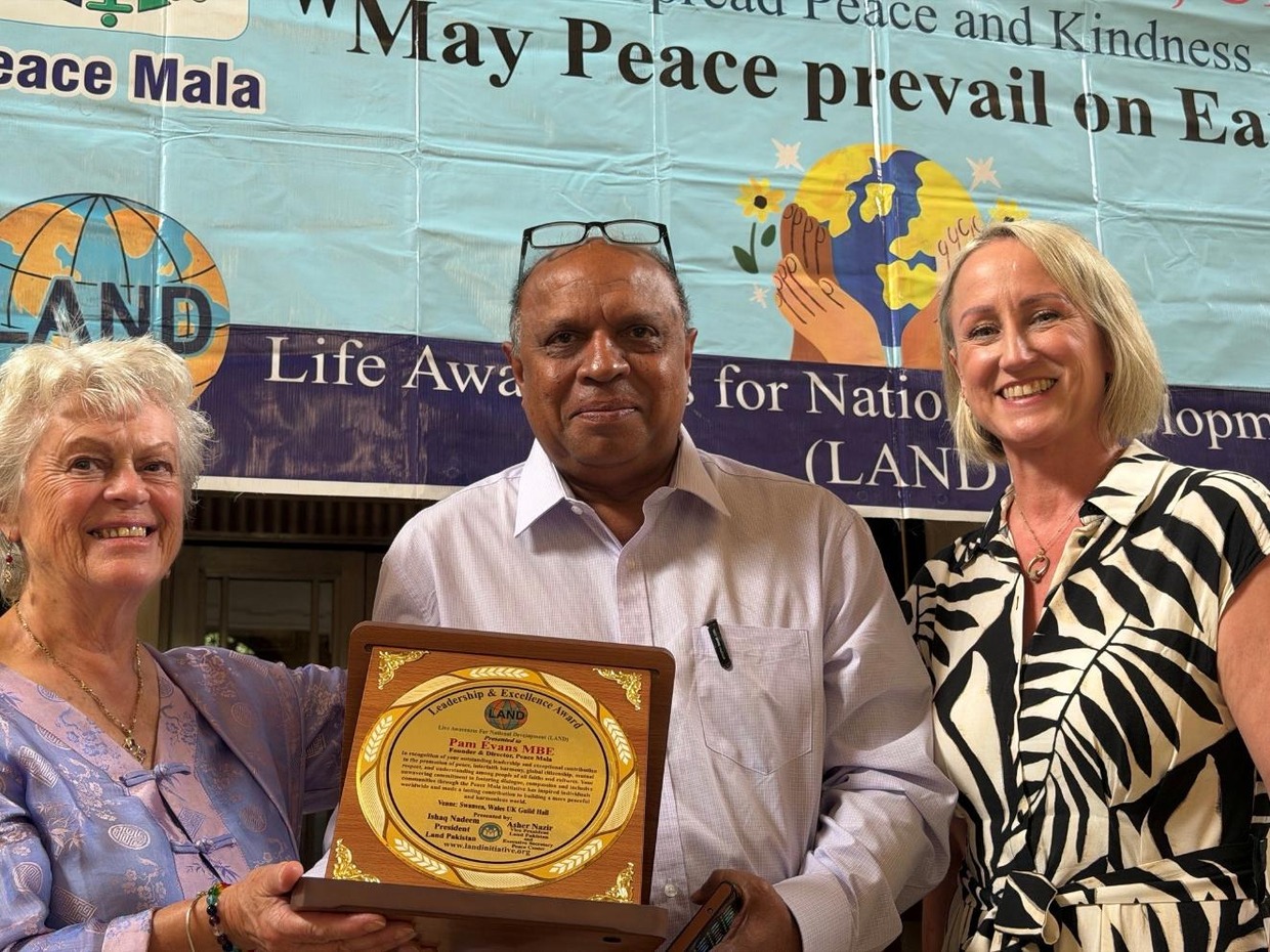 Pam Evans MBE, Mubarak Ali and Cllr Andrea Harrington holding the Leadership and Excellence Award plaque in front of a Peace Mala banner