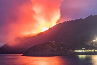 Orange smoke rising from a burning hillside above the lit shopfronts on Pendine seafront