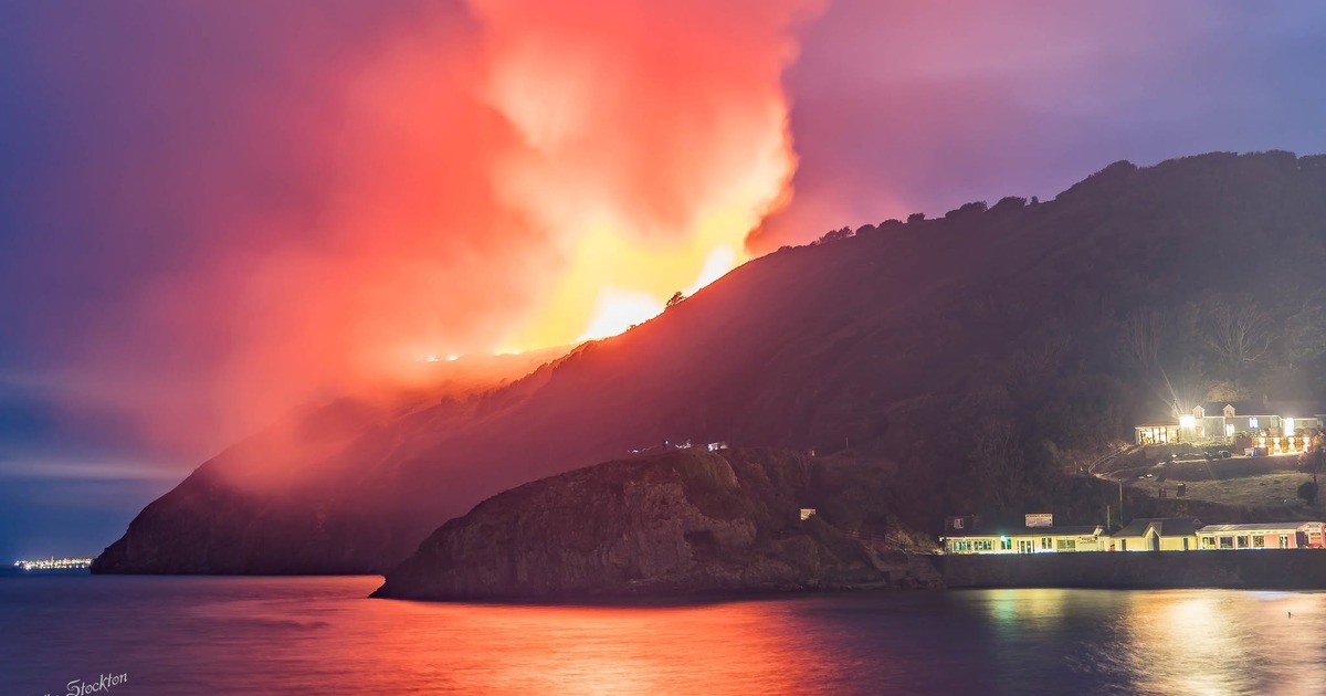 Orange smoke rising from a burning hillside above the lit shopfronts on Pendine seafront