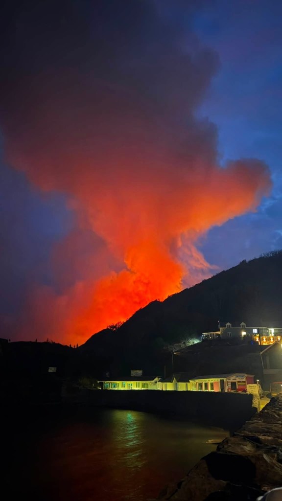 Red smoke lit by fire on a dark hillside above lit seafront shops at Pendine