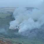 Smoke drifting across fields and woodland in daylight after a wildfire near Pendine