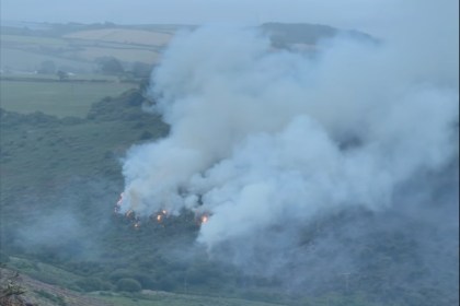 Smoke drifting across fields and woodland in daylight after a wildfire near Pendine