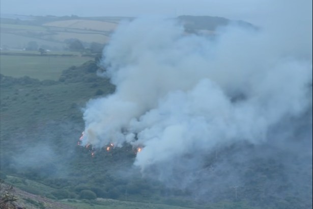 Smoke drifting across fields and woodland in daylight after a wildfire near Pendine