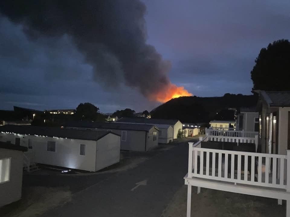 A column of dark smoke and orange flame on a hillside beyond rows of static caravans