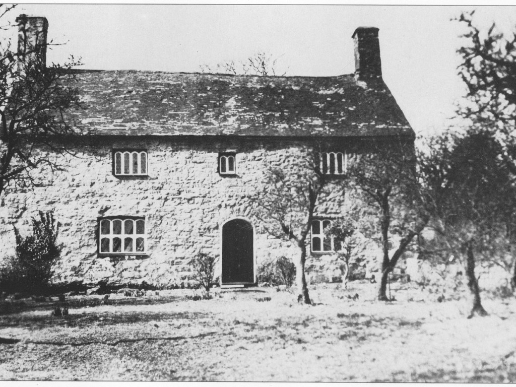 Black and white photograph of a long stone farmhouse with mullioned windows and two chimneys behind bare trees