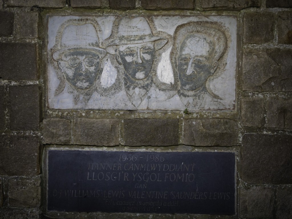 Carved stone panel showing the faces of the three men above a slate plaque with a Welsh inscription dated 1936 to 1986