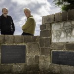 Two men standing behind a stone memorial wall at Penrhos carved with three faces and Welsh inscriptions