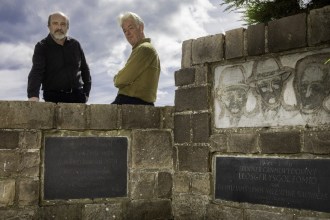Two men standing behind a stone memorial wall at Penrhos carved with three faces and Welsh inscriptions