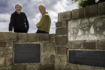 Two men standing behind a stone memorial wall at Penrhos carved with three faces and Welsh inscriptions
